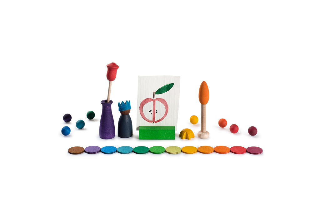 The image shows colorful wooden toys and objects arranged on a white surface. There is a circular wooden board divided into colored sections labeled with the months of the year. In the center, there is a small blue figurine. Surrounding the board, there are various wooden pieces including balls, a bowl with colored balls, a wooden flower in a vase, and dice with numbers. Additionally, there is a cardboard box with illustrations and a painting of a person under a rainbow.