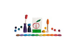 The image shows a collection of colorful wooden toys and objects arranged in a visually appealing manner. The objects include various shapes such as spheres, discs, and figures, all painted in vibrant colors. In the center, there is a small card with a drawing of a peach, held upright by a green stand. The arrangement of the objects and the use of bright colors make the image interesting and visually engaging.