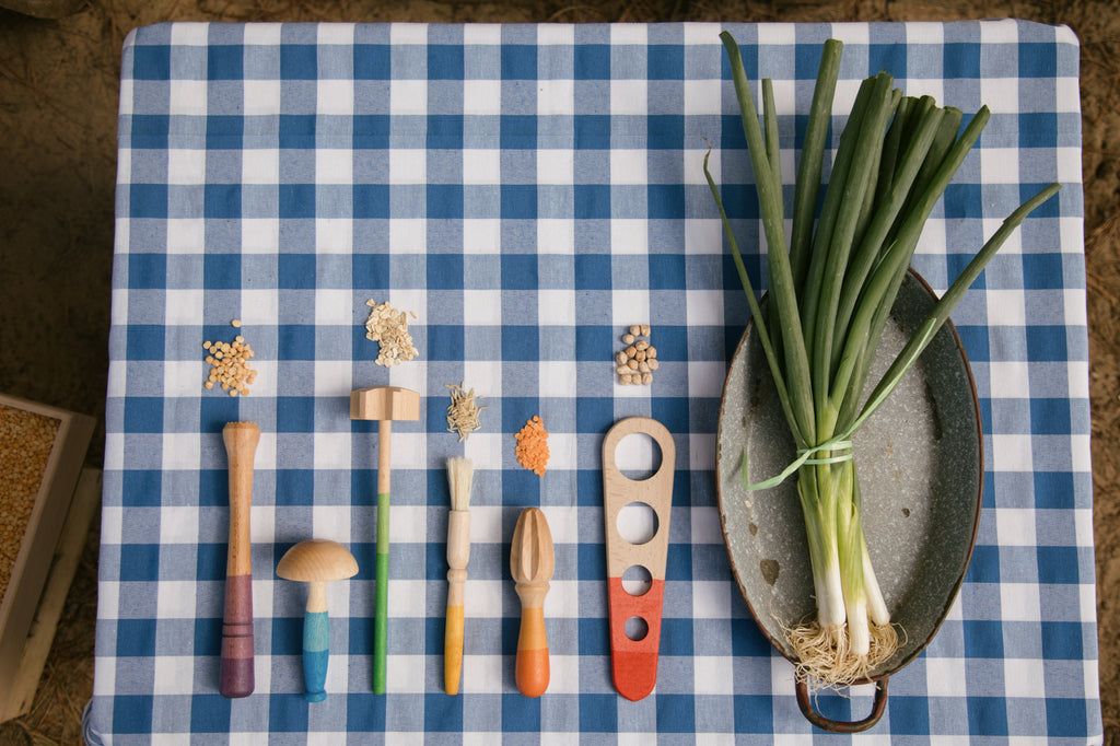 The image shows six wooden kitchen utensils arranged in a row. Each utensil has a distinct shape and a portion of each handle is painted in a different color. From left to right, the utensils are:

A wooden spaghetti measure with three circular holes and a red-painted handle.

A wooden citrus reamer with an orange-painted handle.

A wooden pastry brush with a yellow-painted handle.

A wooden meat tenderizer with a green-painted handle.

A wooden mushroom-shaped tool with a blue-painted handle.

A wooden mud