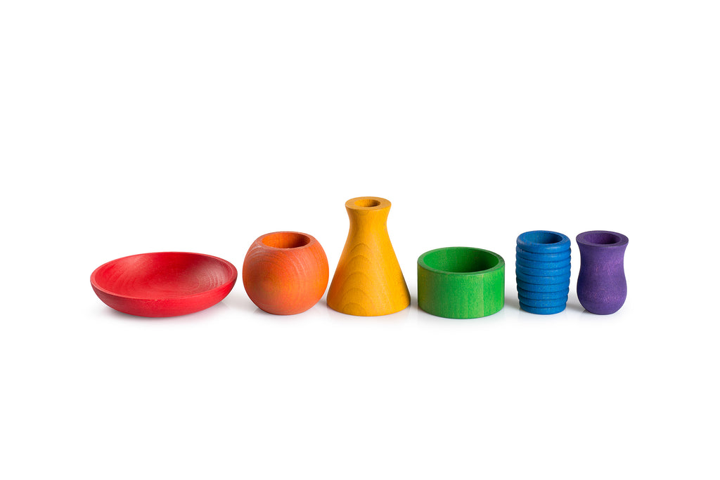 A collection of colorful wooden objects is displayed on a white background. The objects include a red bowl, an orange rounded vase, a yellow conical vase, a green cylindrical container, a blue ribbed vase, and a purple small vase. Each object is uniquely shaped and brightly colored, showcasing a variety of forms and hues.