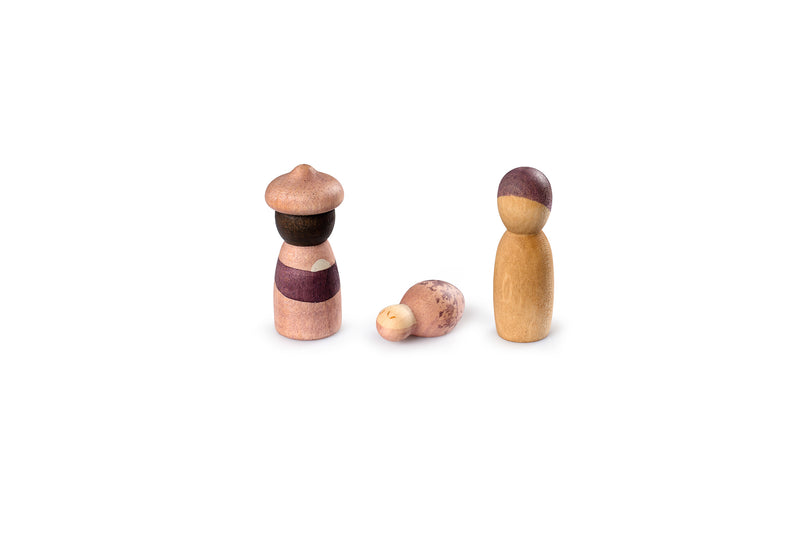 The image shows three wooden figurines. One figurine is standing on the left, wearing a hat and a striped outfit. The second figurine is lying down in the middle, resembling a baby. The third figurine is standing on the right, with a simple design and a two-tone color scheme. The figurines are arranged against a plain white background.
