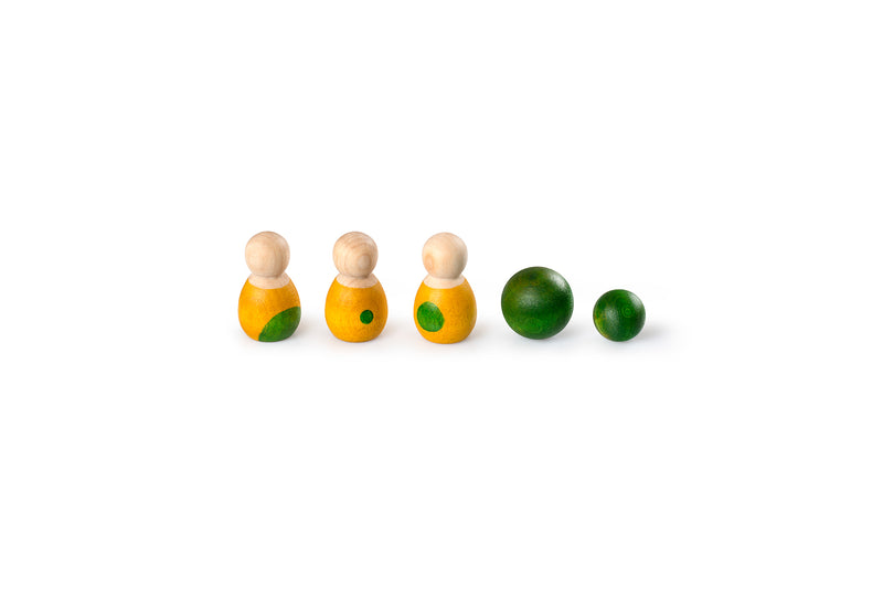 The image shows four wooden figurines with natural wood heads and yellow bodies accented in green, and two green wooden balls of different sizes. The objects are arranged in a straight line, with the figurines on the left and the balls on the right. The toys are simple yet colorful, designed for educational or play purposes.