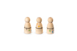 The image shows three wooden peg dolls with round heads and cylindrical bodies. Each doll has colorful decorations: green and blue dots with a ladder and house drawing on the first, orange and yellow dots with a house and tree on the second, and green, blue, and purple dots with a flower on the third. The dolls are arranged in a row and have a playful appearance.