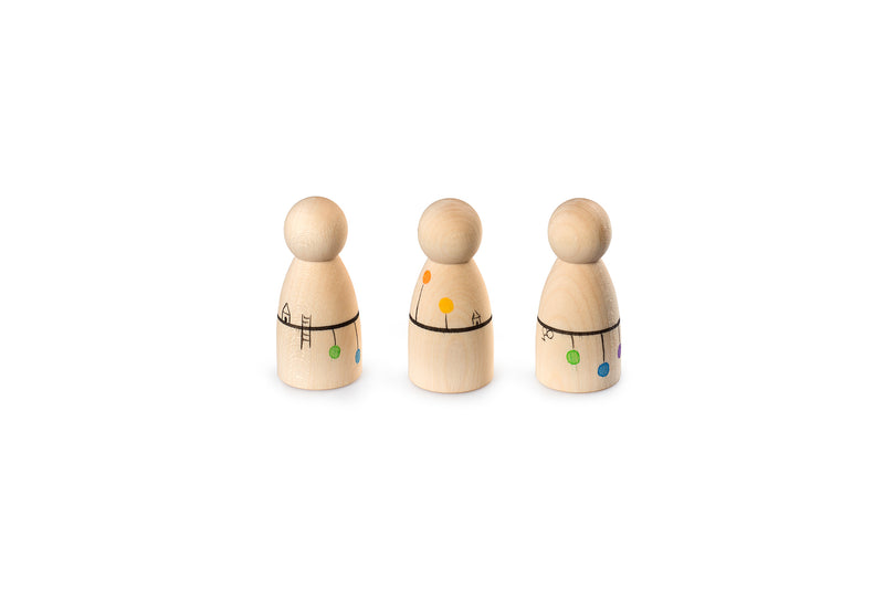 The image shows three wooden peg dolls with round heads and cylindrical bodies. Each doll has colorful decorations: green and blue dots with a ladder and house drawing on the first, orange and yellow dots with a house and tree on the second, and green, blue, and purple dots with a flower on the third. The dolls are arranged in a row and have a playful appearance.