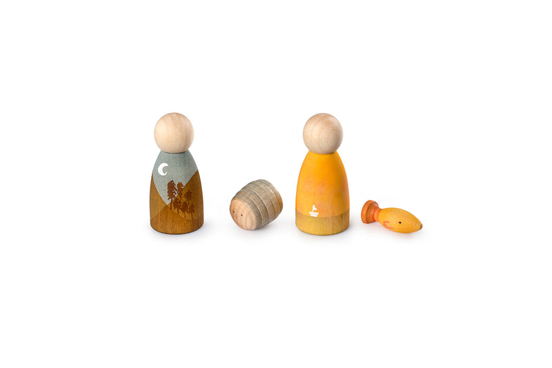 The image shows four wooden toys arranged in a row. From left to right, there is a wooden figure painted with a landscape of trees and a crescent moon, a small cylindrical wooden piece, another figure painted yellow with a white arrow, and a small wooden fish. The toys are simple and colorful, likely designed for children’s play.