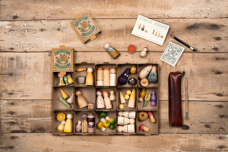 The image shows a cardboard box with multiple compartments, each holding a variety of small, colorful wooden figurines and objects. The figurines come in various shapes and sizes, resembling people, animals, and abstract forms. Inside the box, there are also two smaller boxes labeled "LUCKY," featuring a design with a bird and a banner. Additionally, there is a pencil case with a pencil and a pen, along with a couple of instruction cards displaying illustrations of the figurines.