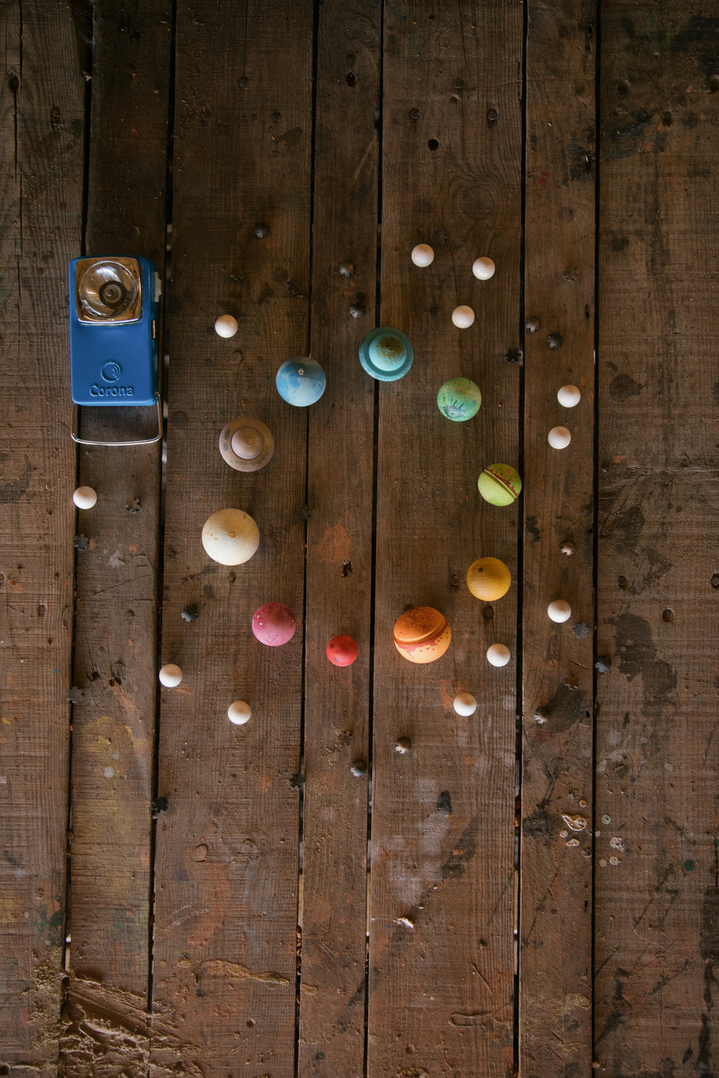 A circular arrangement of colorful wooden balls and star-shaped pieces on a white background. The balls are painted in various colors and patterns, resembling planets and celestial bodies, with some in blue, green, yellow, orange, red, and brown tones. Smaller beige wooden balls and dark brown star shapes surround the larger spheres, creating a look reminiscent of a solar system with orbiting elements