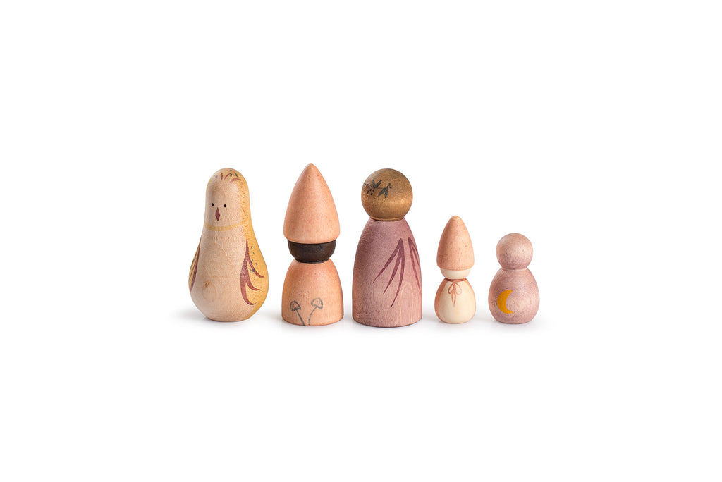 An assortment of small, handcrafted wooden figures arranged in front of a cardboard box with black, nature-inspired artwork. The figures include various shapes and colors resembling mushrooms, cones, and abstract human forms, each painted in soft, earthy tones like green, pink, and beige. The box design shows a whimsical scene of plants, stars, and an owl on a branch against a black background, evoking a night-time, nature-inspired theme.
