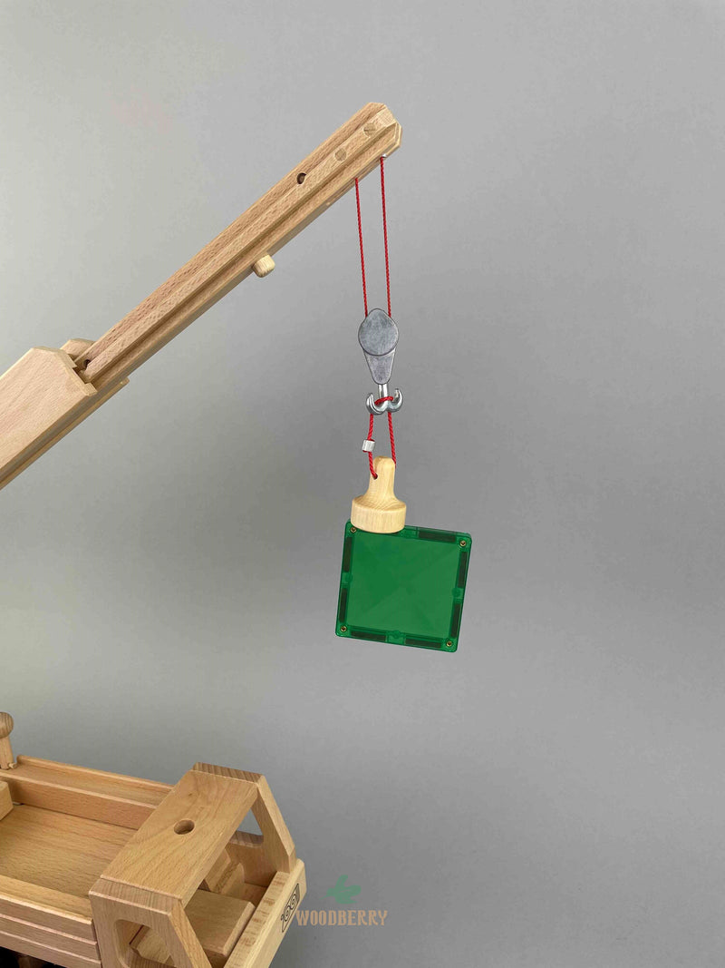 A wooden lifting magnet with red loop hanging rope by Fagus toy. A green 1x1 size Connetix magnetic tile is being lifted by the Fagus lifting magnet. All hanging on the extended arm of the Fagus mobile crane to demonstrate the magnet strength