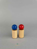 Two wooden peg dolls standing next to each other. The blue one on the left and the red one on the right. They have ball shape heads and natural wooden color cylinder as bodies.
