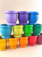 A set of 12 wooden mates, or toy cups, in rainbow colors and stacked in a display. Five cups on the bottom row (dark red to orange), 4 in the middle row (green to light blue), and three on the top row (blue to violet). Image is closeup and at an angle.