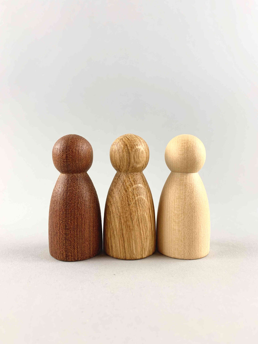 Three wooden peg figure toys in three different natural wood tones from dark to light, placed on wooden blocks in the shape of stairs. 