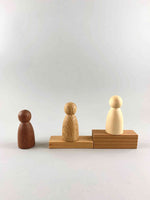 Three wooden peg figure toys in three different natural wood tones from dark to light, placed on wooden blocks in the shape of stairs. 