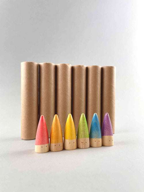 Wooden mini rainbow colored baby sticks by Grapat toys in Spain. The pointed top small peg people sticks line up in a row. From left to right is red, orange, yellow, green, blue and purple. Six standing cardboard tubes in the background