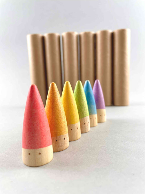 Wooden baby sticks. Six rainbow colored pointed top little peg creatures with two hand painted eyes lining in a row from close to far. With six cardboard tubes in the background.