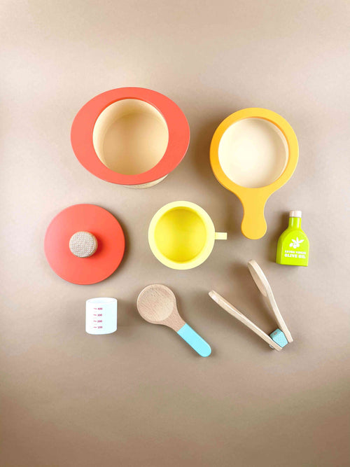 Wooden toy for pretend play and kitchen play. Wooden stock pot in red,  wooden frying pan toy in yellow. Wooden measuring cup toy in white. Wooden tong. Wooden olive oil toy. Wooden yellow mug toy. Wooden toy spoon with blue handle. By Woody Puddy Japan